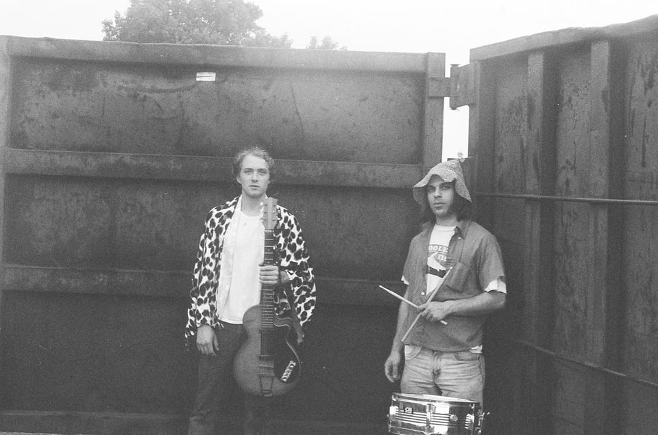 A man in a leopard-print jacket holding a guitar upright stands next to a man in a floppy hat holding crossed drumsticks in his left hand. They are flanked by the walls of an industrial-sized dumpster.