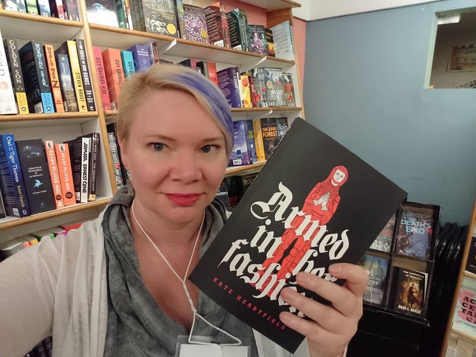 Kate with a blue strak in her hair and headphones in her ears, holding a copy of Armed in Her Fashion near a bookshelf.