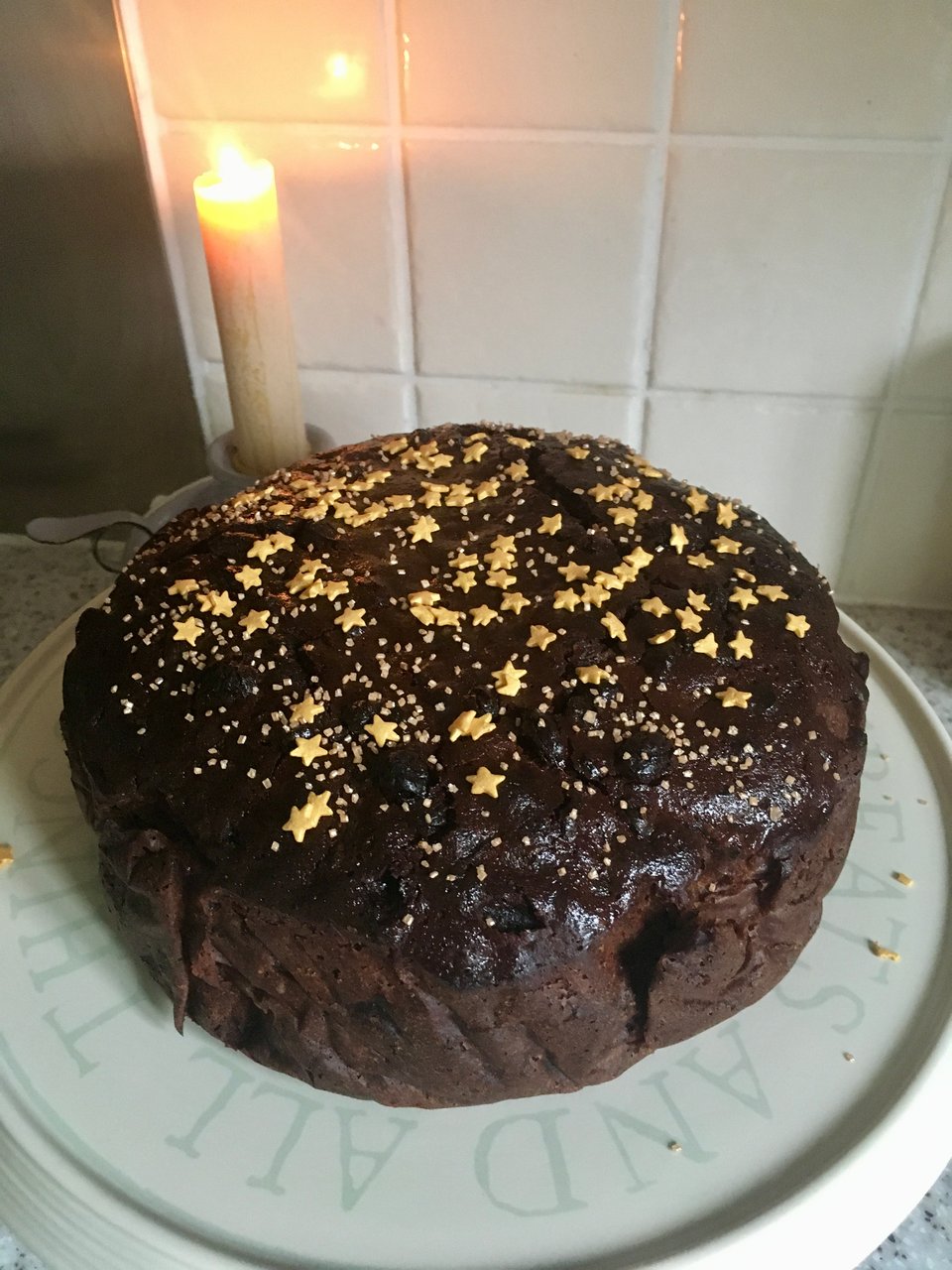 A dark brown cake on a ceramic stand is scattered with gold stars and sugar sprinkles. There's a tall beeswax candle illuminating the photo.