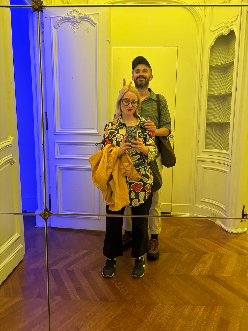 A selfie of me and Todd in an old wall-length mirror. He's giving me bunny ears. The mirror is lit yellow and blue from the exhibitions on either side of the vestibule.