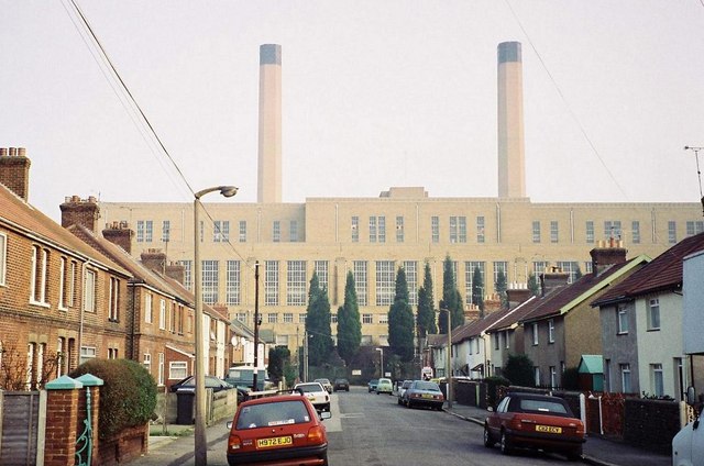 A street with two storey Edwardian brick terraces on one side, and 1960s council semis on the other. At the end of the street, rising way above the houses and a line of trees, is a huge brick building with tall windows and two very tall chimneys above them.