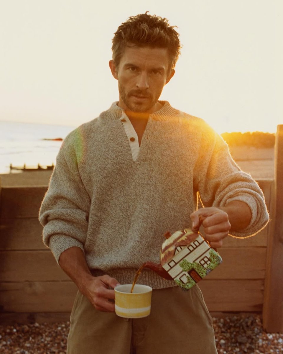Photo from a photoshoot of actor Jonathan Bailey. He stands on a beach wearing a grey sweater and pouring himself a cup from a house-shaped teapot whilst staring into the camera.
