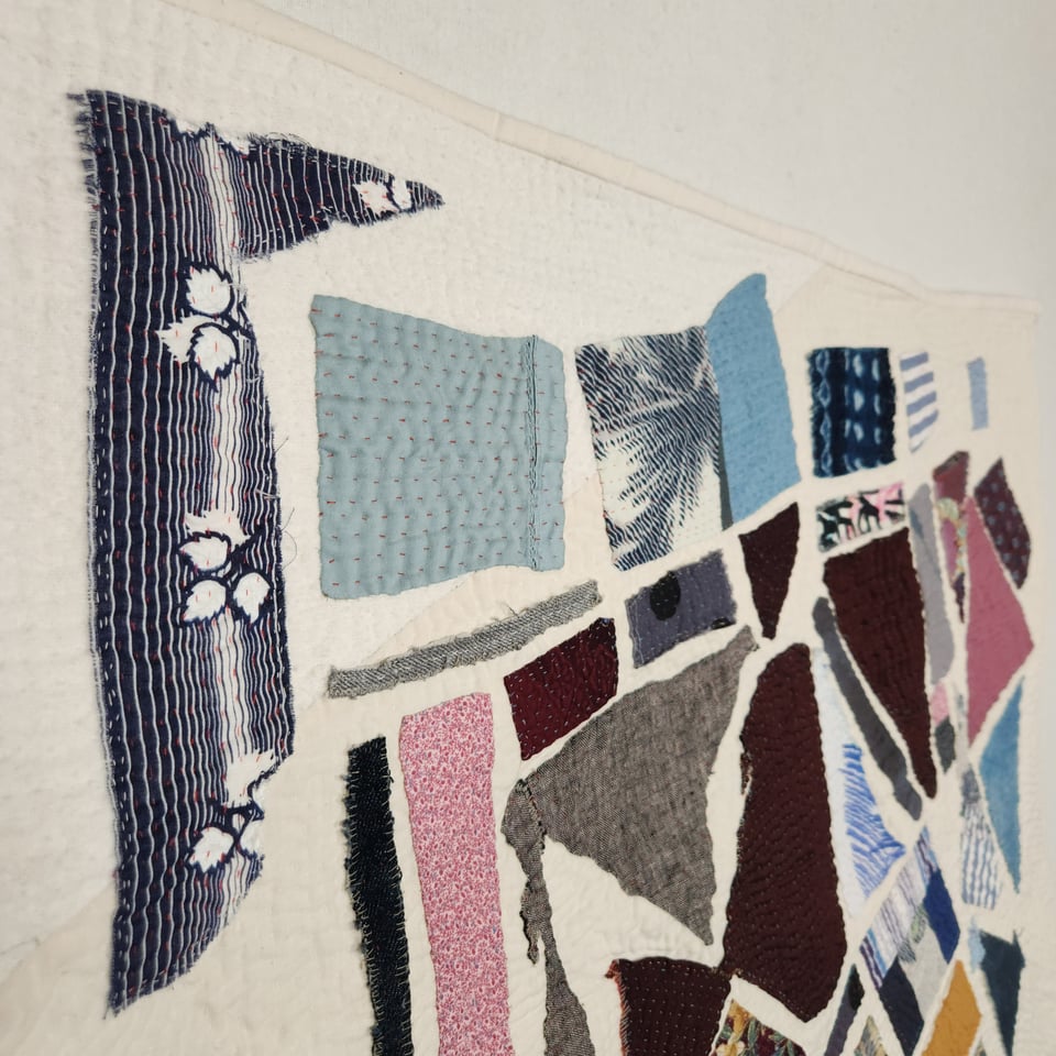 Side view of Meet Me at the Corner, a patchwork quilt by Sarah Atlee. In the foreground is a jagged scrap of fabric with white stripes and leaves on a navy blue background. In the rest of the photo other scraps and hand stitches are visible.