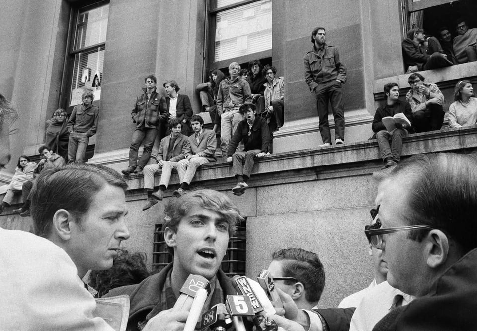 A young man is interviewed with microphones in the foreground, while other young people sit and stand on various parts of the architecture behind him.