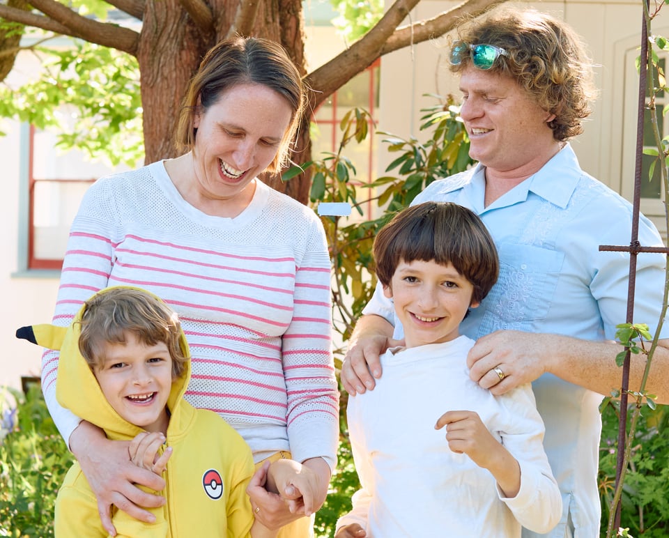 My family stands in a sunny garden, dressed informally. Me and my wife are in the back, similing down on our children. The kids, ages 6 and 9, face the camera, laughing.