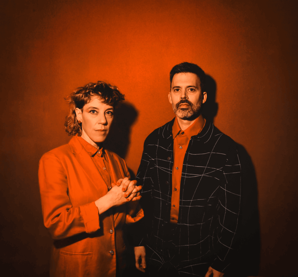 A woman with short, curly hair wearing a red blazer and red button-up shirt stands next to a man with a short beard in a red button-up shirt and black, patterned jacket. They are in front of a red background.