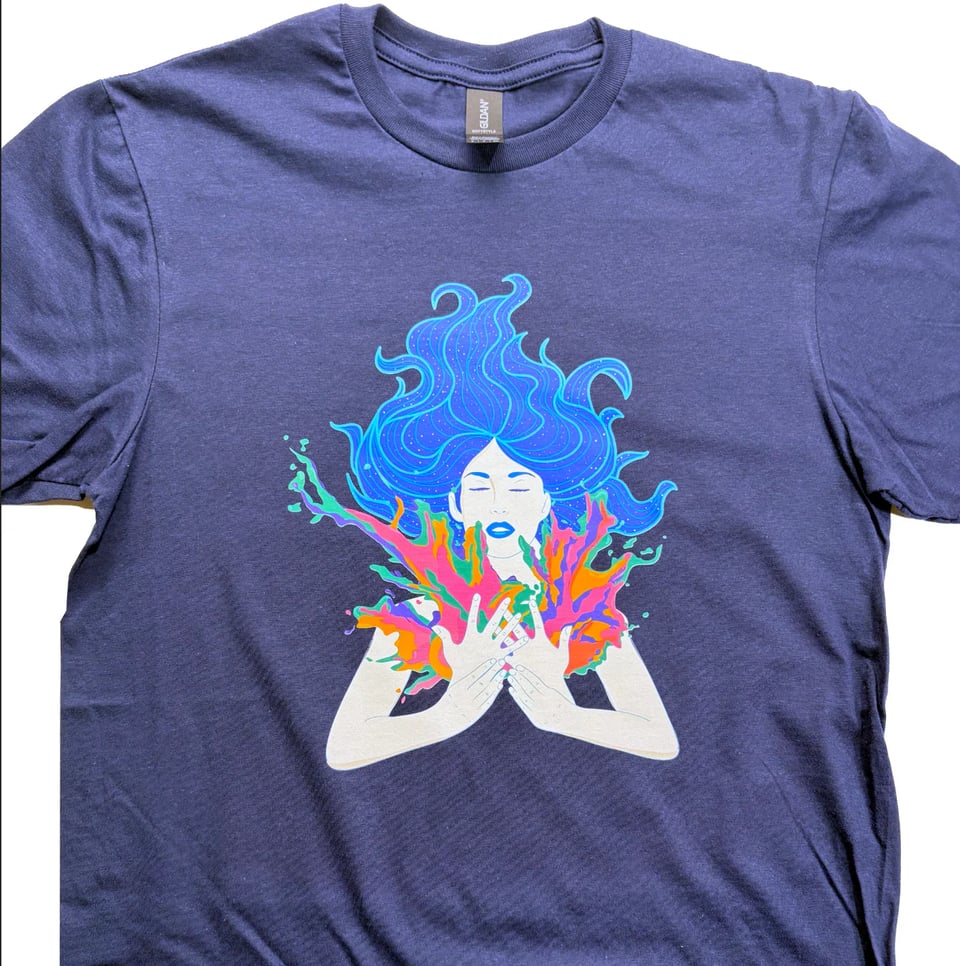 Commercial image of a t-shirt on a blank background. On the front of the blue shirt is the image of a woman raising her hands in front of her body, the ASL sign for INSPIRE. From her hands spill multicolored magic energy, and her long blu hair streams out from the top of her head with the force of the inspiration.