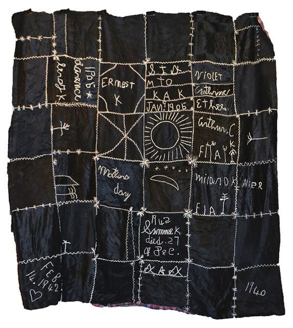 A black quilt with white stitching. There are words and signatures and little line drawings embroidered.
