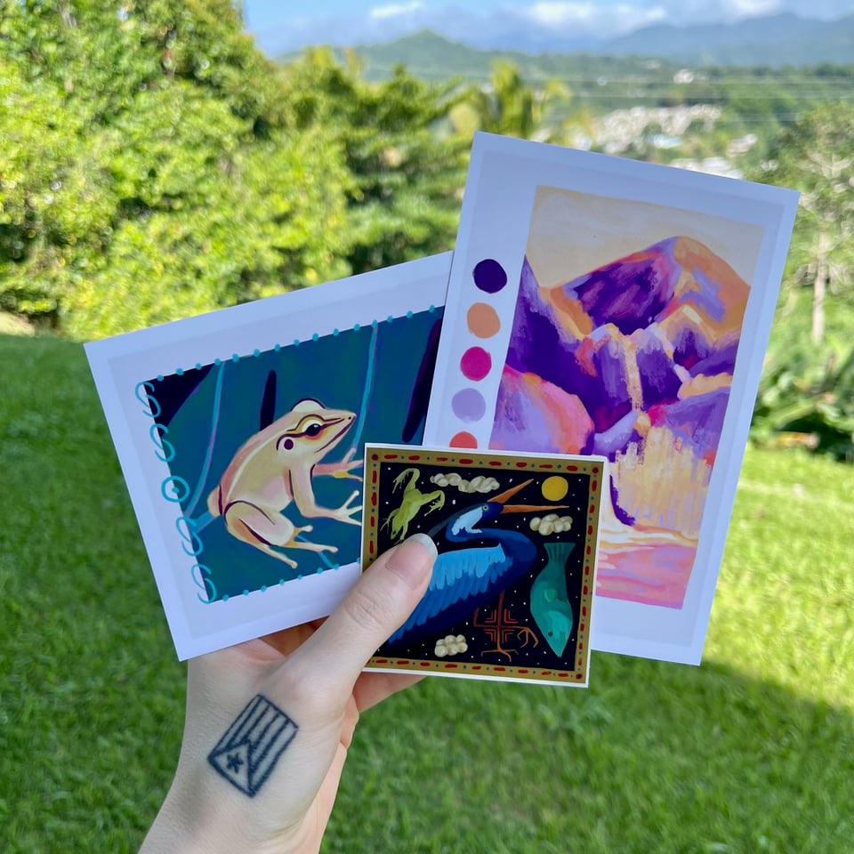 Photo of 2 colorful postcards and a sticker, background is a Puerto Rican landscape