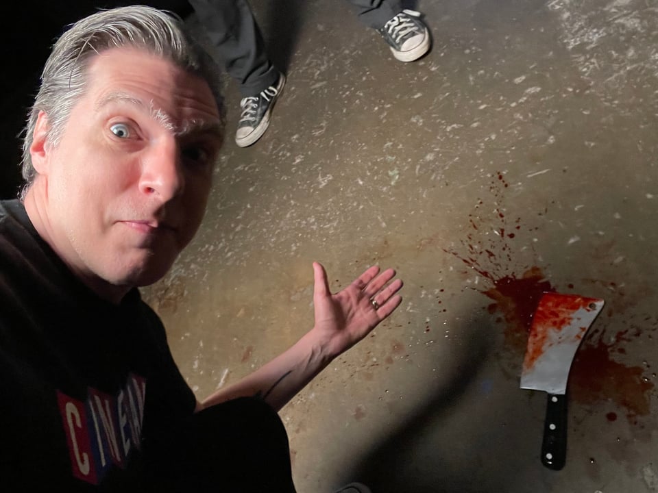 A man leans down and points, smiling, at a prop cleaver on the floor in a pool of splattered blood