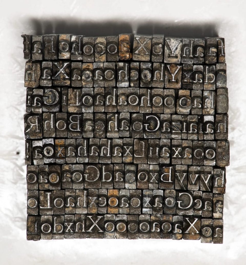 A photo of a grid of letters done in metal type. There are arranged with about 20 letters across and eight down, forming a cube. The letters are reversed to our view, and they do not spell anything; they seem to be randomly arranged. The metal type letters look very old and slightly corroded.