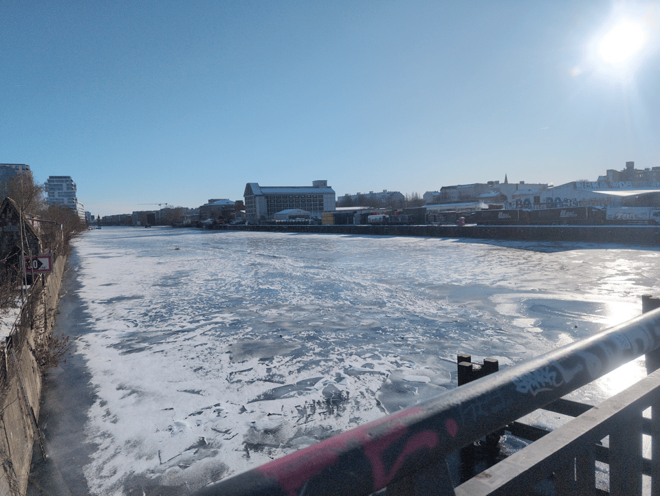 The Spree River is frozen. The ice isn't smooth, and it looks like several chunks of ice have flowed from upstream and gotten stuck on the bridge, where they have re-frozen. The sky is a clear, light blue, and the sun is in the top right corner, casting a reflection on the ice below.