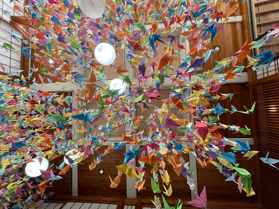 Hundreds of colorful origami cranes strung on strings hanging from the ceiling. The picture is shot from below looking up at the cranes from underneath.