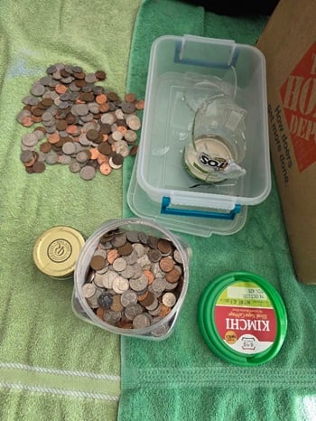 Multiple items on green towels: a pile of assorted coins, a plastic kimchi bucket half-full of assorted coins and lid, a four ounce glass jar of just quarters with a gold lid, a rectangular plastic bin with a broken glass salsa jar and bits of glass.