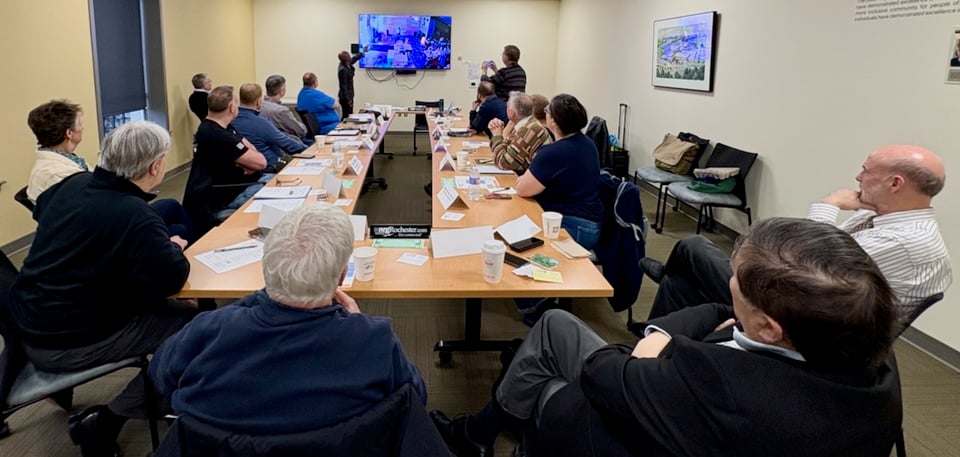 Shantae is motioning to a TV screen showing surveillance video from inside a business during a robbery to a group of people seated around tables set up in a long hollow rectangle.