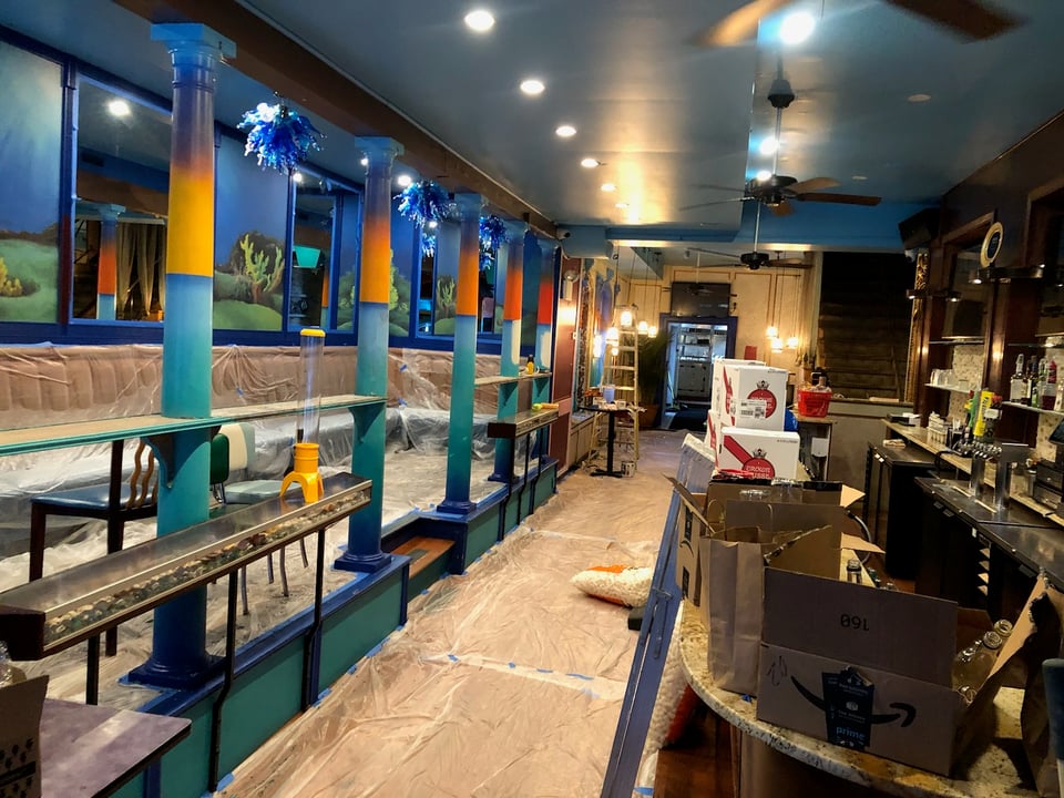 Image of the bar from the front door, with an island sunset ombre on the pillars lining the gallery seating, and light sandy back panels, and a bright blue ceiling.