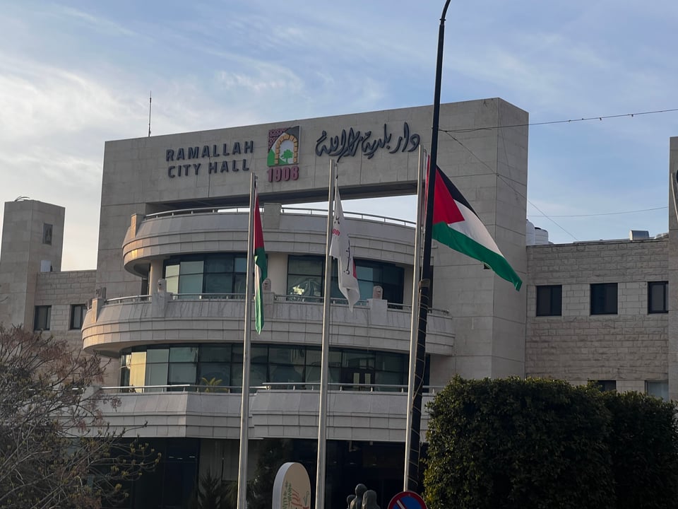 A 3-story modern building that says Ramallah City Hall, and text in Arabic, with Palestinian flags in front