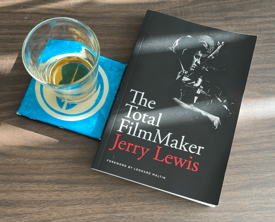 Sunlight hits a table with a filmmaking book by Jerry Lewis and a glass of whiskey