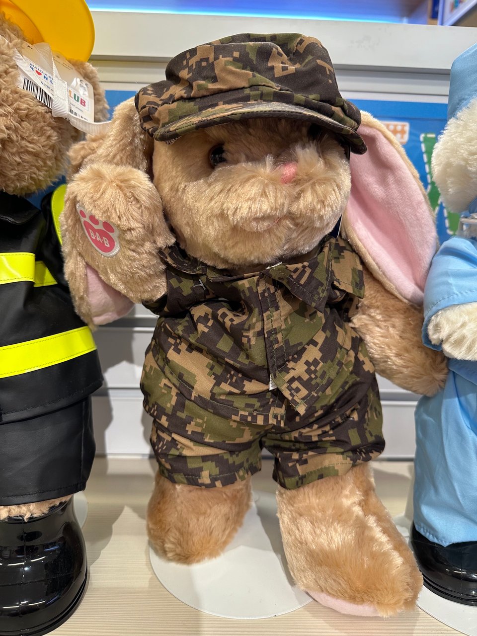 A rabbit soft toy with the Build-a-Bear logo on its paw. It is wearing a camouflage uniform with cap. The camouflage pattern consists of blocky, pixellated areas of green and brown.
