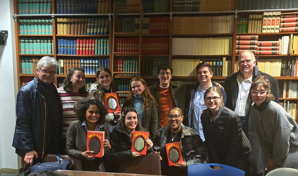 A dozen adults, comprising a faculty member, me, and students, pose in two rows against a library wall full of books, smiling. Four of us are holding up copies of HUMANS.
