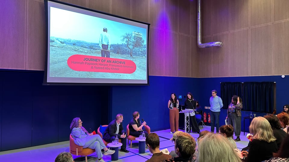 Photograph of various people on a stage in front of an audience. Three presenters and an MC are standing behind a lectern to the right of the stage as we look, and they are being questioned by a panel of three seated people in the centre. Above the stage is a large screen with a still from a film and a caption which says, "JOURNEY OF AN ARCHIVE: Hannah Papacek Harper, Francesco Amoruso & Yazeed Abu Khdeir"