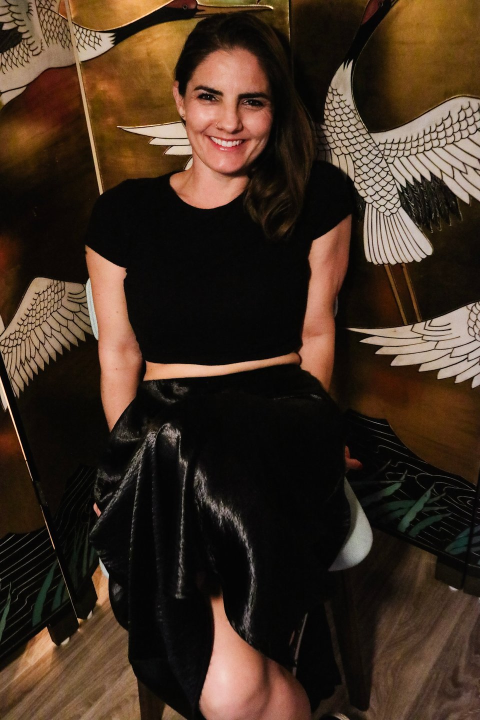 A smiling woman (Ariana) is sitting on a chair against a gold background with birds, leaned back with her hands down by her sides.
