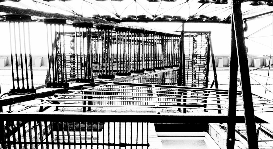A black and white photo of a fire escape on the side of an apartment building, taken from the ground looking up. The steps and walkways are in contrast to a washed out bright white background.