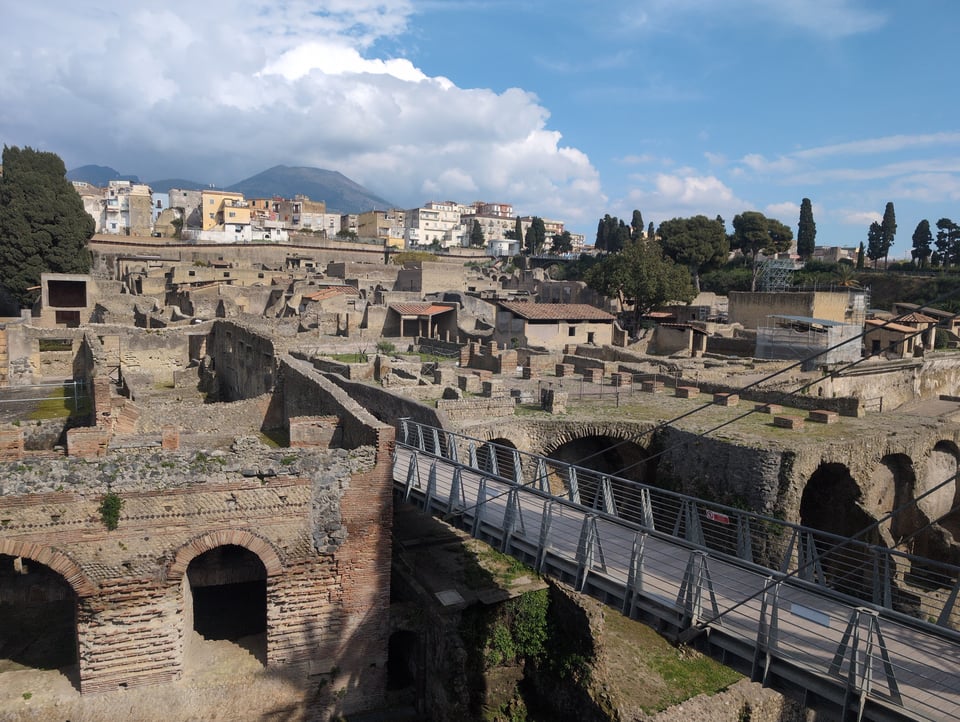 In the foreground is a picture of the excavation site at Herculaneum. It is mostly broken stone and brick buildings, with a bridge leading from off screen to the site. In the middle ground is the modern city of Ercolano, above the tops of the ruined buildings. There is a brick wall at the back edge that stabilizes and separates the ancient city from the modern. In the distant background is Mount Vesuvius, wreathed in storm clouds. The rest of the sky is sunny.

I was definitely a normal child of the 1980s mutually assured destruction era who had no negative effects of it on her psyche.