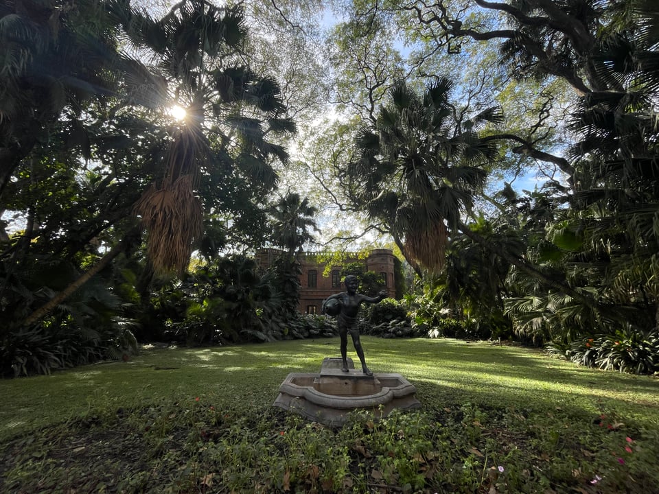A small statue in the middle of lawn which is surrounded by a variety of tropical trees. In the background is the main house of the Botanical Garden.