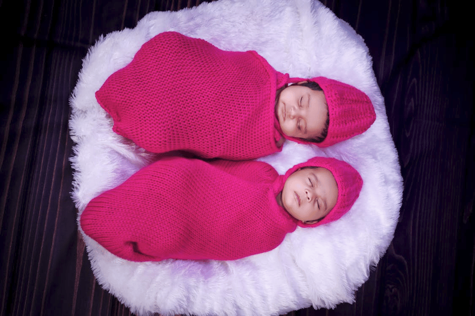 A photo looking down on two infants sleeping. They're both dressed in bright knitted outfits that swaddle their bodies and have caps over their heads. They are lying on a white fluffy round rug