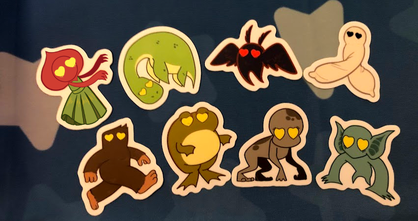 A photo of 8 cryptid stickers lined up next to each other: the flatwoods monster, the loch ness monster, mothman, fresno nightcrawler, bigfoot, loveland frog, dover demon, and hopkinsville goblin.