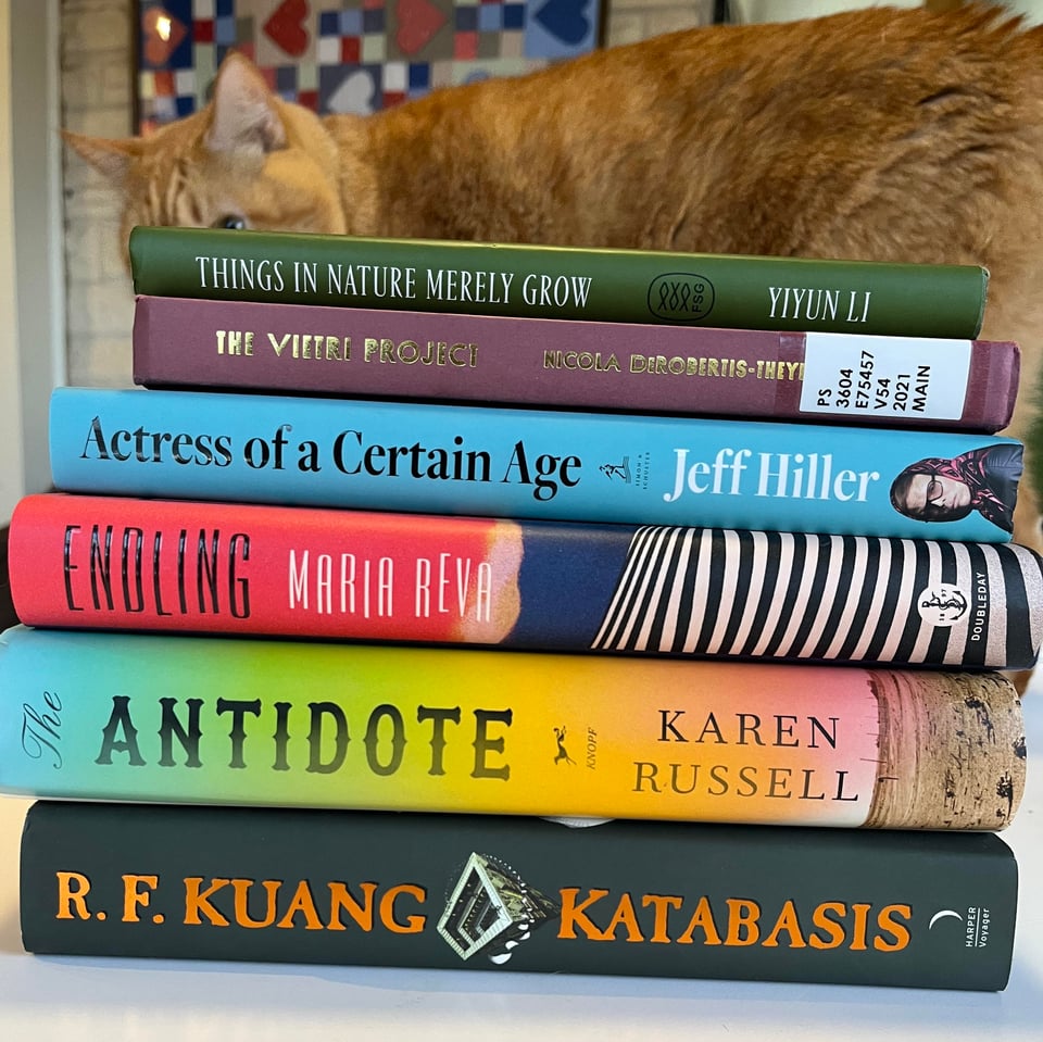 A stack of books on a table with an orange tabby cat sniffing behind them