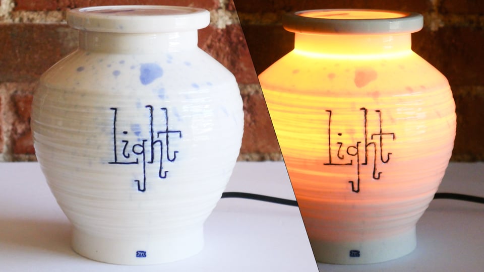 white porcelain jar in split view, glowing amber from the inside when turned on. From the base it flares out and up like a honey pot, and then angles back in near the top, to an opening that matches the base. The side is textured in overlapping grooved rings, and light is carved in an upright sort of slab cursive. The lid is on and also glowing from inside. There's a splatter of subtle blue wash from above.