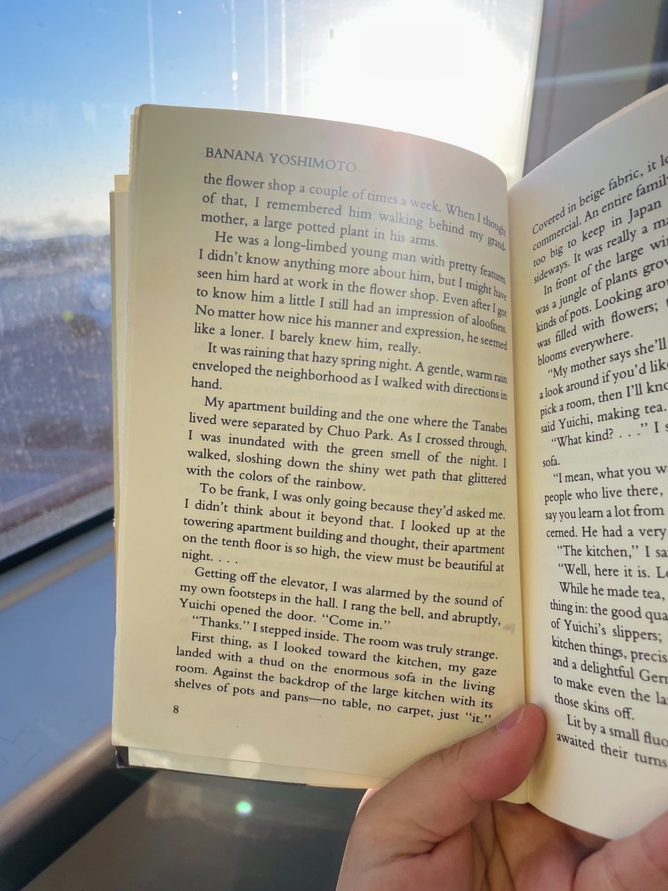 A photo of an open book, taken by someone riding a train at golden hour, sun glare flaring. The book is "Kitchen" by Banana Yoshimoto.