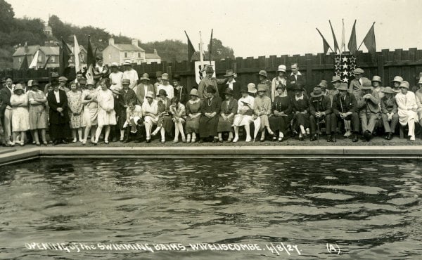 A postcard showing the opening of Wivey pool in 1927. Two rows of chairs have been set up, and older men and women are sat looking at the empty water. At one end, a lot of younger women are standing, many with their arms folded. There are a lot of hats being worn.