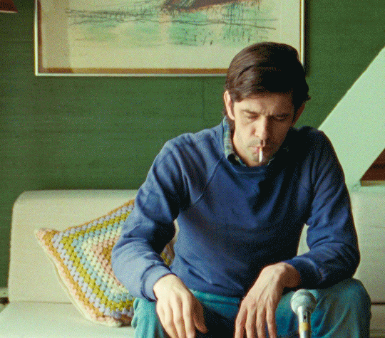 Ben Whishaw as Peter Hujar sitting on a cream-colored couch smoking a cigarette and shaking his head