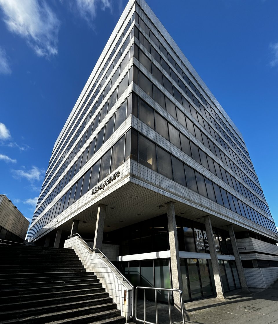 A square building sitting on top of a glass podium and thin concrete piloti. The building looks like a layer cake, with ribbons of glass and tiling all the way around. In the foreground, some concrete steps rise up to meet the main square building.