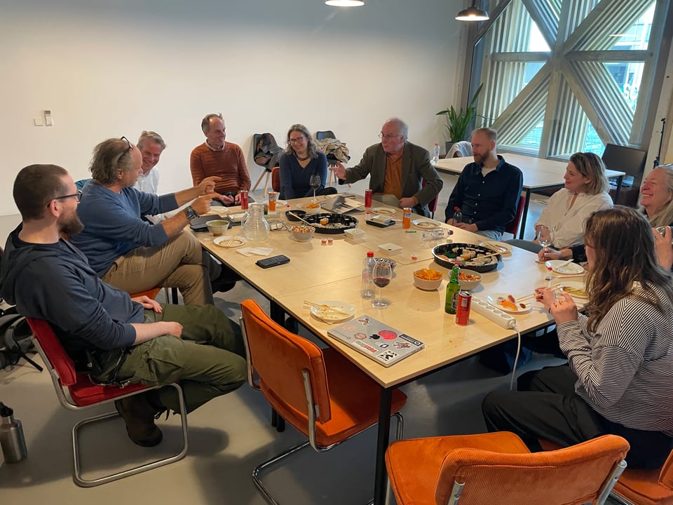 A group of people sit around two tables with drinks and sushi. Two men gesticulate in conversation, most other people are laughing.