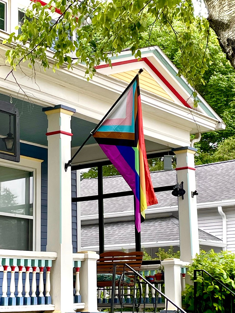 A Progress Pride flag is flying on a black flag pole from one of my house's porch columns. The body of the house is painted dark blue with white trim and yellow, teal, and red accents.