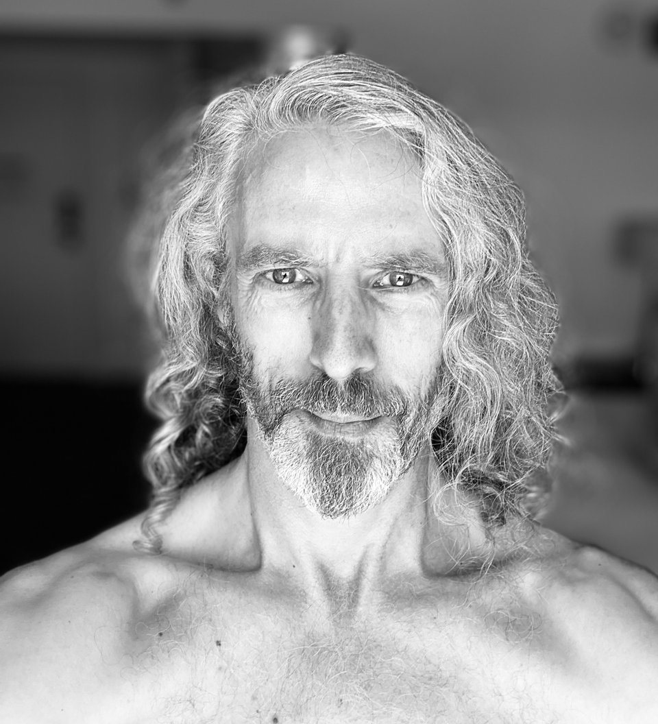 B/W photo of a shirtless man, from mid-chest up. He has long hair and a trim beard, and in the bright light he looks like the Silver Surfer's skeevy cousin