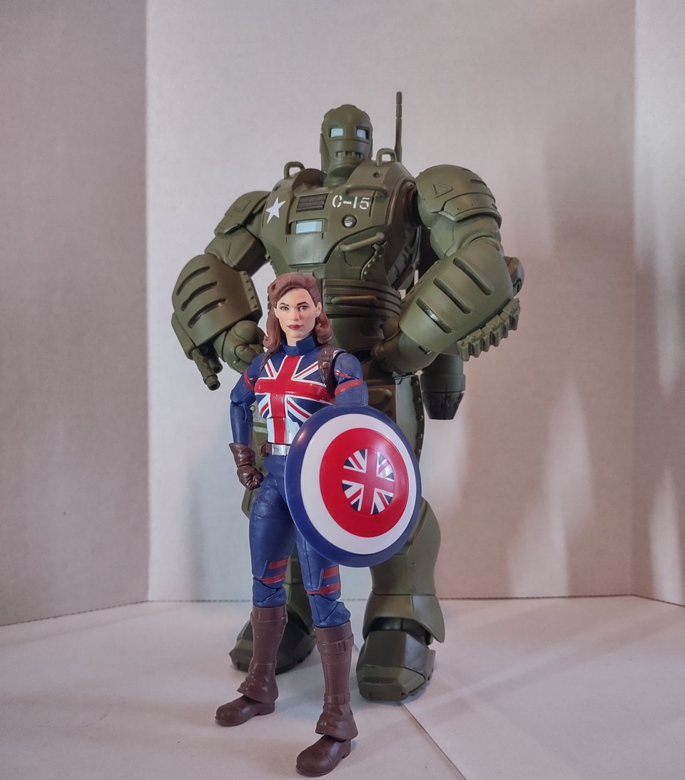 Two action figures in a white photo booth. In the foreground is Captain Carter- the alternate-world Peggy Carter who got the super-soldier formula- standing with her shield. Looming behind her, almost twice her height, is the Hydra-Stomper, an olive-drab armored suit that looks like a WWII-tech Iron Man.