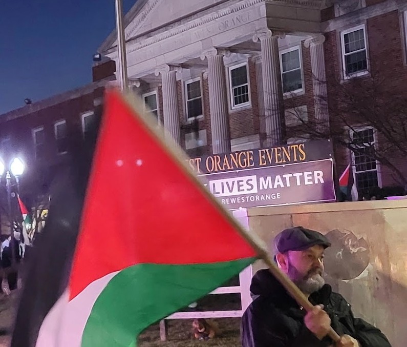 A man holds a Palestinian flag in front of the "Black Lives Matter" sign in front of West Orange Town Hall. It gives the effect of giving the message "Palestinian Lives Matter"
