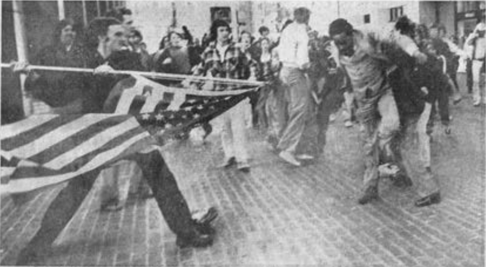 A black and white photograph of a Black man in a plaza being attacked by a White teenager brandishing the American flag.