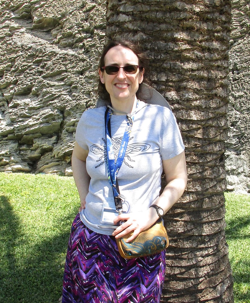 The author in a grey shirt, purple skirt, leaning against a palm tree in the sunshine.