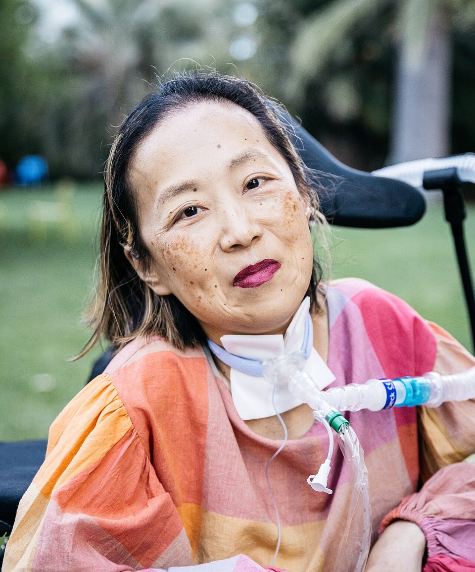 A smiling freckled woman in a wheelchair with dark hair, purple lipstick, tracheostomy tube and patterned blouse