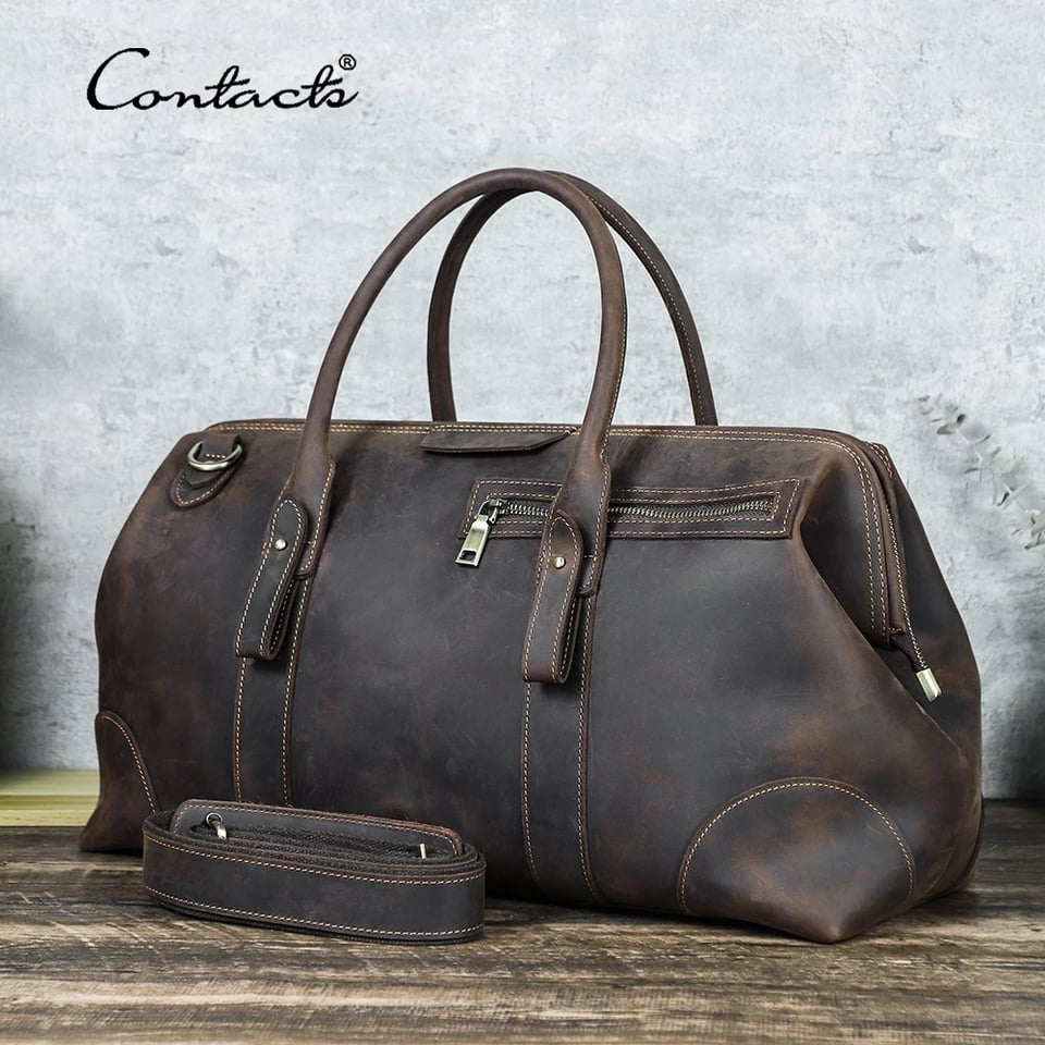 A picture of a leather carryall/duffel bag, coffe brown, with two handles and a longer strap, sitting on a wooden surface. The brand name (Contacts) is in the top left corner.