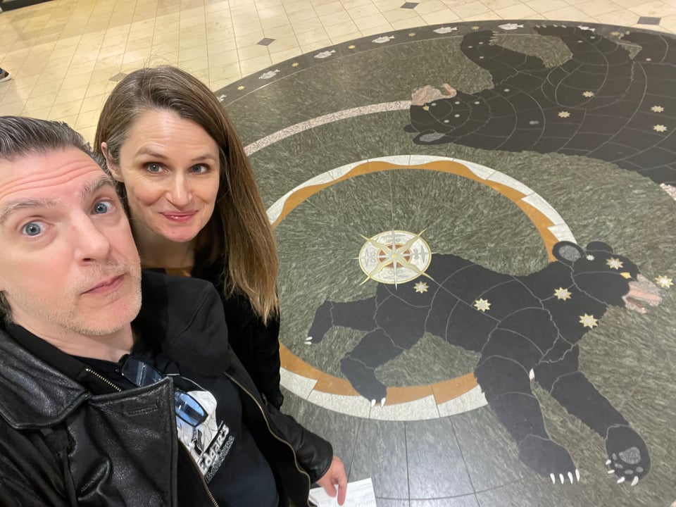 A man and a woman smile at the camera, on the floor beneath them is artwork of the Ursa Minor constellation aka a blitzed out bear rubbing its butt on stars