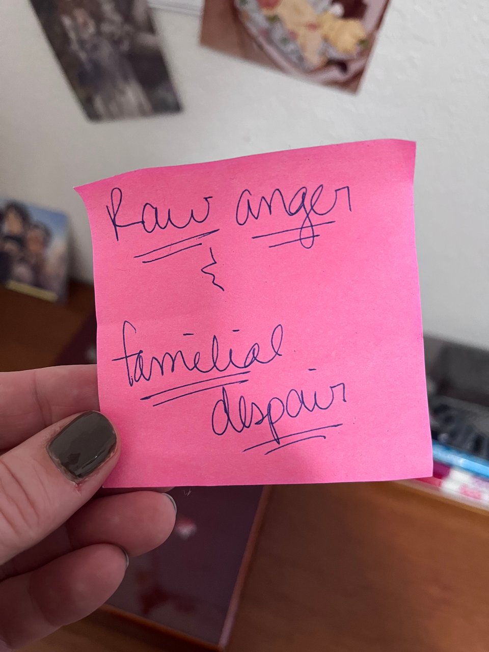 hand with gray nail polish holding up a neon pink sticky note that reads in messy handwriting "raw anger & familial despair" against a blurry background of a whiteboard and shelf