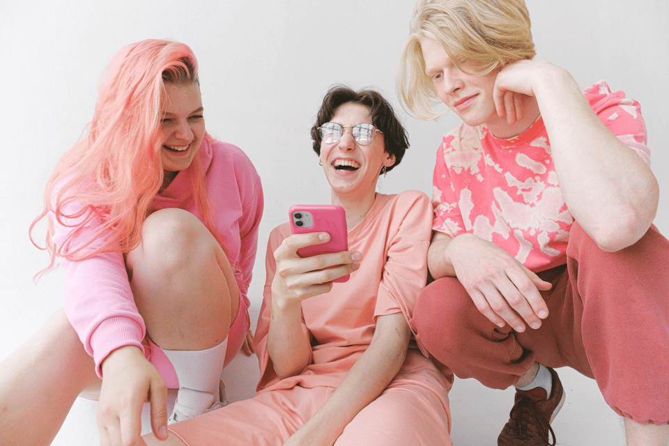 A picture of three people crouched down on the ground, two of whom are smiling, and laughing, while the other one merely smiles. They're all dressed in bright, pink, and the one on the far left has very long flowing brightly dyed pink hair