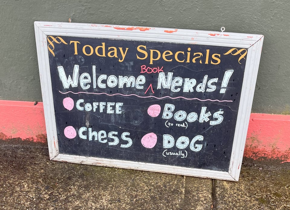 A photograph of a chalkboard sign outside of Sam's Cara café. The frame is light blue. The text at the top reads "Today Specials". Written in chalk is, "Welcome ^Book Nerds!" In bullet points underneath, it reads: "Coffee, Chess, Books (to read), Dog (usually)."